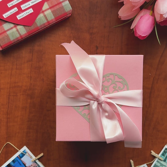 Video demo of a pink birthday explosion gift box: the lid lifts and the box’s sides tumble outward to reveal three handcrafted layers — a four-photo grid at the top, origami wallet pockets holding small prints, and inner flaps with handwritten messages. Close-ups show pearl-finish paper, crisp archival prints, and a secret pocket that produces a heartfelt surprise.