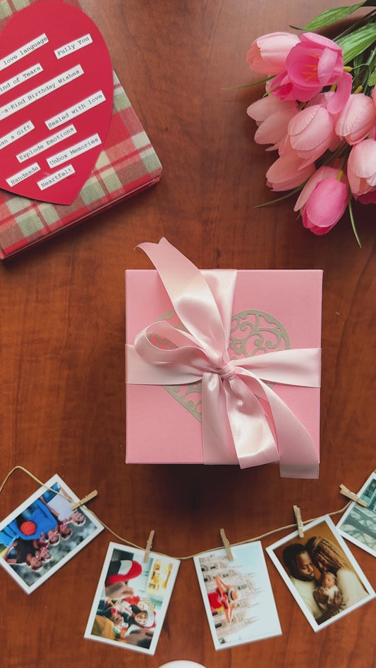 Video demo of a pink birthday explosion gift box: the lid lifts and the box’s sides tumble outward to reveal three handcrafted layers — a four-photo grid at the top, origami wallet pockets holding small prints, and inner flaps with handwritten messages. Close-ups show pearl-finish paper, crisp archival prints, and a secret pocket that produces a heartfelt surprise.